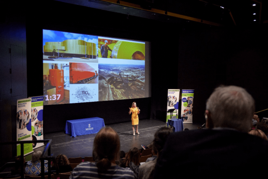 An image from the Swansea University 3MT final. A presenter stands on a large stage wearing a yellow dress, in front of a large slide depicting images of painted metal. The audience watches on in the foreground.