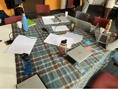 A photograph from inside an space that became an "evaluation cabin", featuring Anne's picnic blanket on a shared work space (a reminder of stepping outside the usual environment), and other objects from the session (laptops, water bottles, written materials, post-its).
