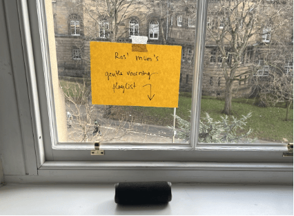 A photograph from inside an "evaluation cabin" space, looking out through a window into a green space, trees, and other buildings. A sign on the inside of the window reads "Ros's mum's gentle morning playlist" and points to a speaker on the windowsill below. In this cabin, we used background music to create a "space that feels different".