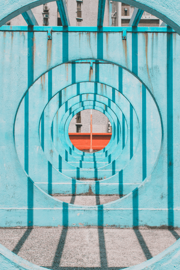 Photograph of a series of concentric turquoise circular frames aligned to create a tunnel-like perspective. Shadows cast by the frames form vertical lines on the ground, with a glimpse of a red horizontal bar and building facade visible through the centre.
